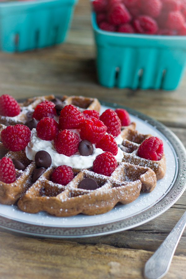 Chocolate Waffles With Fresh Raspberries, topped also with whipped cream and chocolate chips on a plate, with cartons of fresh berries in the background.  