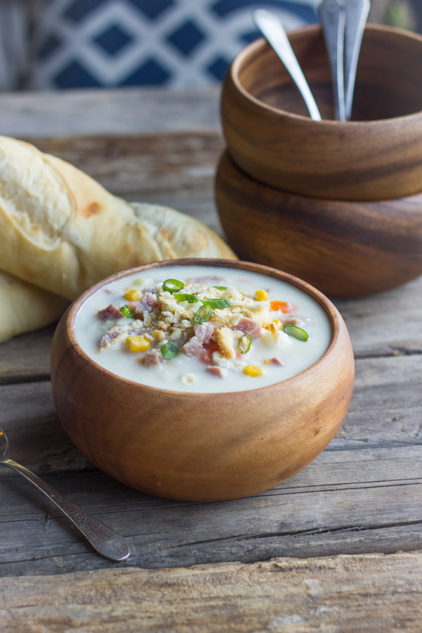 Creamy Potato Soup With Ham in a bowl, with loaves of bread and bowls in the background.  
