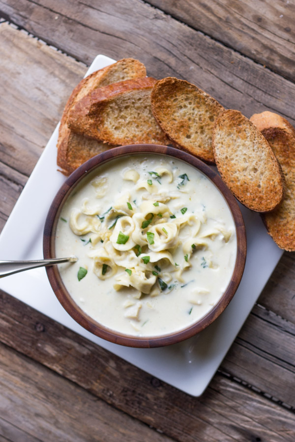 Creamy Spinach And Artichoke Soup With Cheese Tortellini in a bowl that is on a plate with toasted French bread slices.