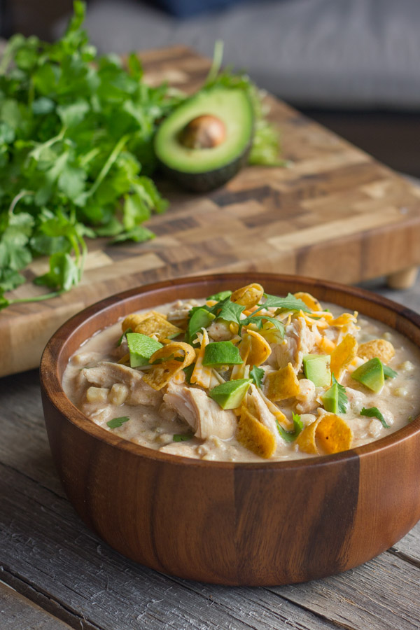 Creamy Crockpot White Chicken Chili in a bowl topped with Fritos, shredded cheese, avocado, and cilantro. There is also a cutting board with cilantro and half of an avocado in the background. 
