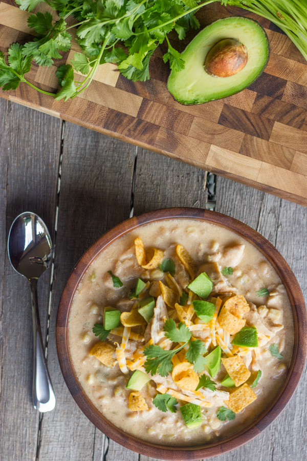Creamy Crockpot White Chicken Chili in a bowl topped with Fritos, shredded cheese, avocado, and cilantro. There is a spoon next to the bowl, as well as a cutting board with cilantro and half of an avocado on it. 