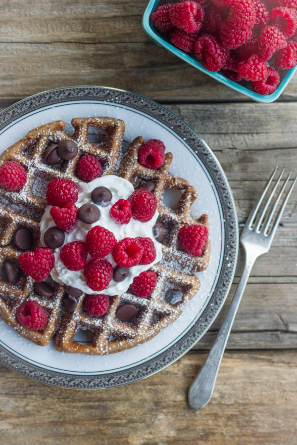 Chocolate Waffles With Fresh Raspberries, topped also with whipped cream and chocolate chips, on a plate with a fork next to it along with a carton of fresh raspberries.
