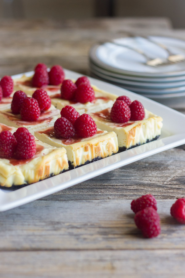 Raspberry Swirl Cheesecake Bars arranged on a serving plate, with a pile of plates in the background with forks on top.  