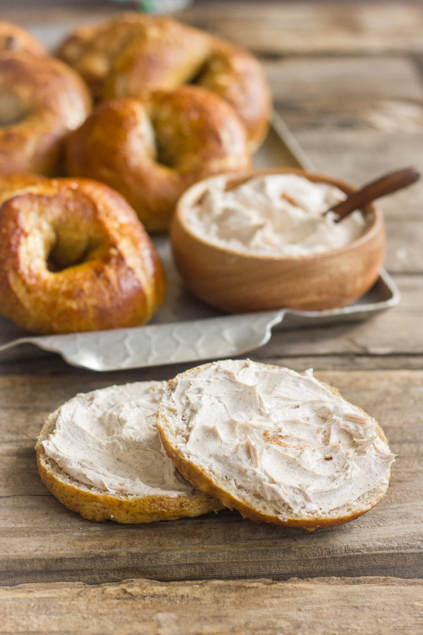 Whole Wheat Soft Pretzel Bagel sliced in half with Cinnamon Sugar Cream Cheese spread on it, and a serving platter of bagels and cream cheese in the background.  