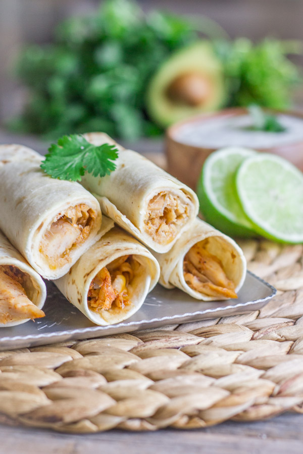 Baked Honey Lime Chicken Taquitos stacked on a serving platter with a bowl of Cilantro Lime Cream in the background.  