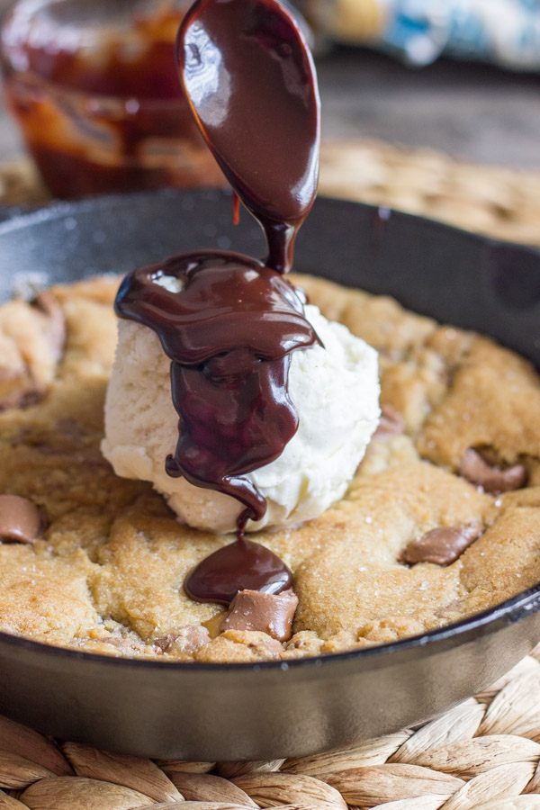 Salted Rolo Chip Pizookie in a small cast iron skillet, topped with vanilla ice cream, and Homemade Hot Fudge being spooned over the ice cream.  