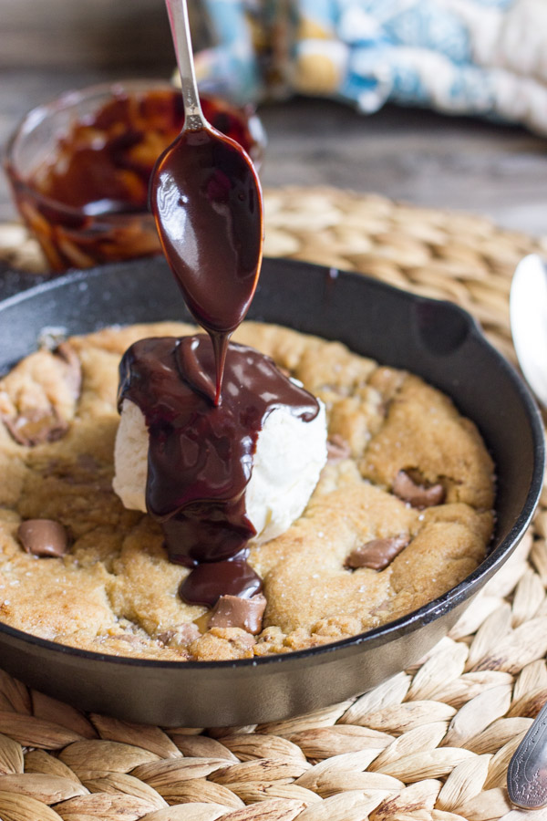 Salted Rolo Chip Pizookie in a small cast iron skillet, topped with vanilla ice cream, and Homemade Hot Fudge being spooned over the ice cream.  