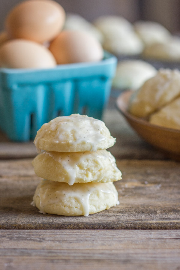 Soft Ricotta Cookies With Vanilla Bean Glaze stacked in a pile of three, with a carton of eggs and more cookies in the background.  