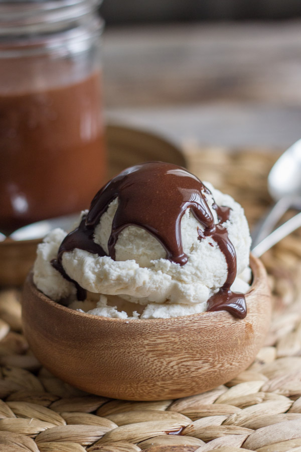 Two Ingredient All Natural Chocolate Syrup on top of vanilla ice cream in a small bowl, with a glass jar of Two Ingredient All Natural Chocolate Syrup in the background.  