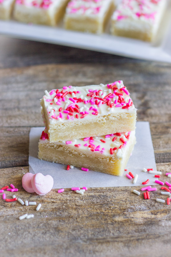 Two Easy White Chocolate Sugar Cookie Bars stacked on top of each other, with sprinkles and two candy hearts near by.  