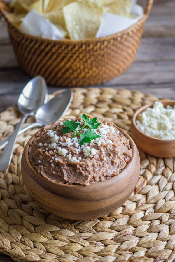 Healthy Crockpot Refried Beans in a bowl topped with queso fresco and cilantro, with a small bowl of queso fresco in the background along with a basket of tortilla chips and two spoons.  