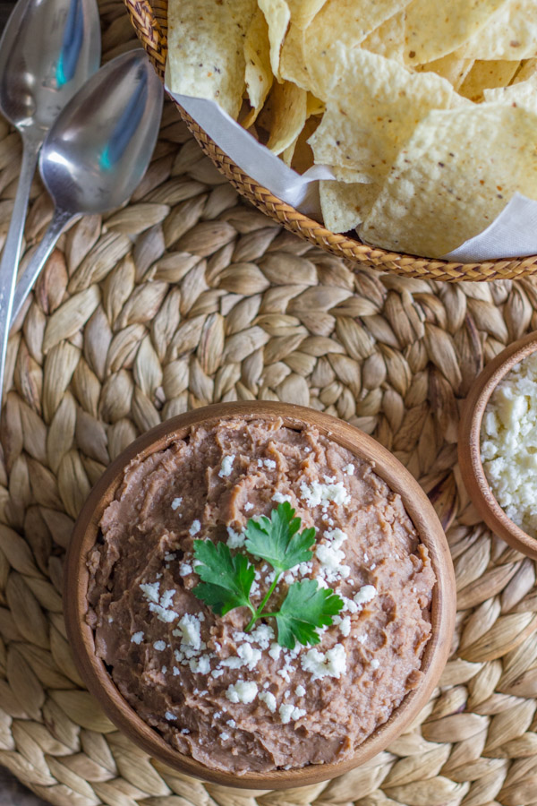 Healthy Crockpot Refried Beans in a bowl topped with queso fresco and cilantro, with a small bowl of queso fresco next to it along with a basket of tortilla chips and two spoons.  
