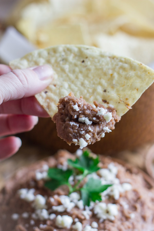 A tortilla chip that has been dipped in a bowl of Healthy Crockpot Refried Beans topped with queso fresco and cilantro.