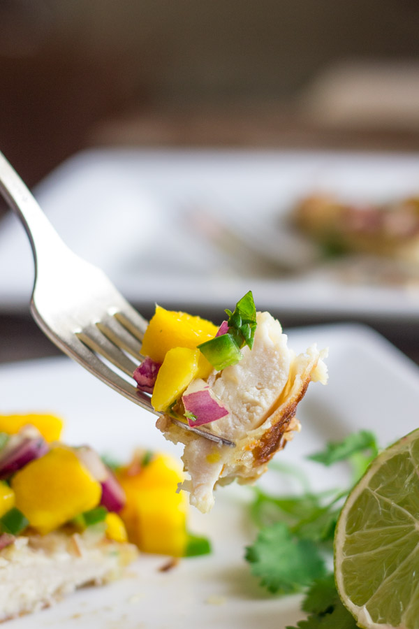 Coconut Crusted Chicken With Mango Salsa bite on a fork with the plated meal in the background.  