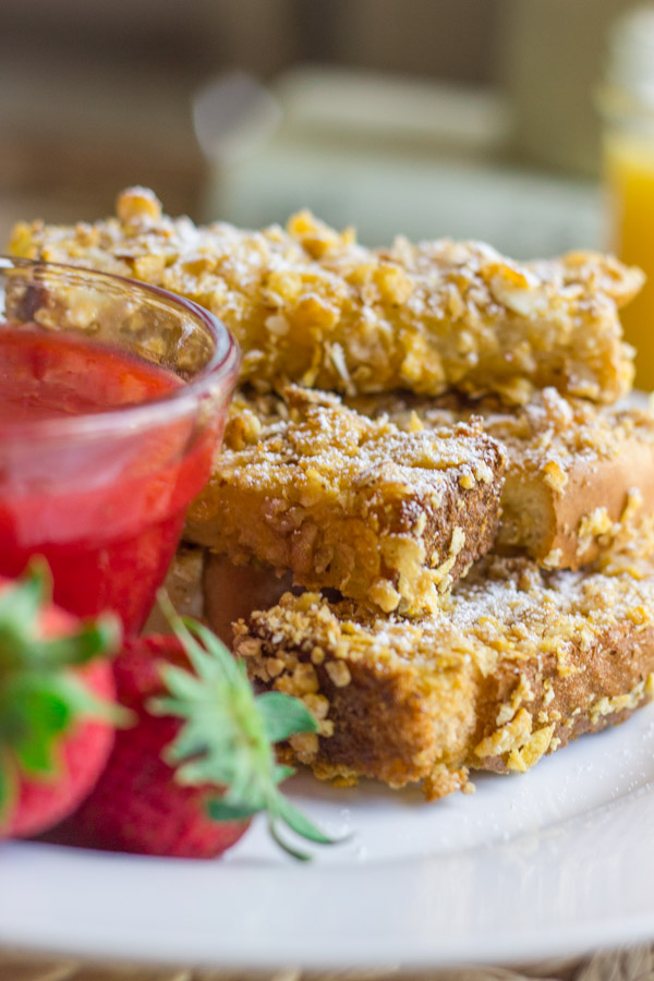 Crunchy Baked French Toast Sticks stacked on a plate next to a glass dish of Strawberry Syrup and some whole strawberries.    