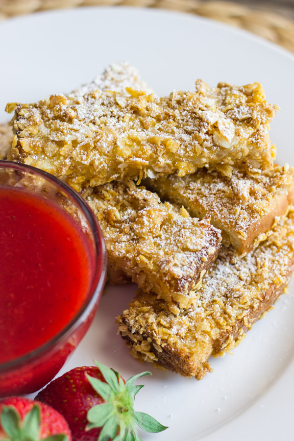 Crunchy Baked French Toast Sticks stacked on a plate next to a glass dish of Strawberry Syrup and some whole strawberries.    