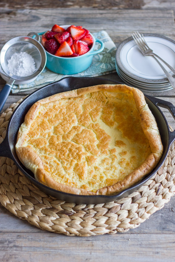 Dutch Baby Pancake in a cast iron skillet, with powdered sugar, a bowl of strawberries and a stack of plates with forks next to it.  