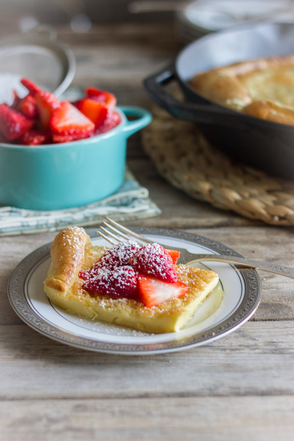 Dutch Baby Pancake slice on a plate topped with strawberries and powdered sugar, with a bowl of strawberries and the Dutch Baby Pancake in a cast iron skillet in the background.  