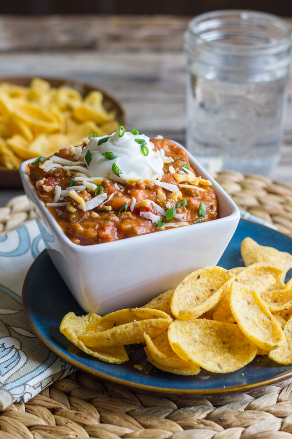 Easy Crockpot Chili in a bowl topped with shredded cheese and sour cream, sitting on a plate with Fritos.  There is a bowl of Fritos and a glass of water in the background.  