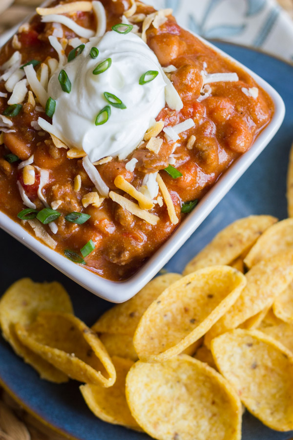 Easy Crockpot Chili in a bowl topped with shredded cheese and sour cream, sitting on a plate with Fritos.  