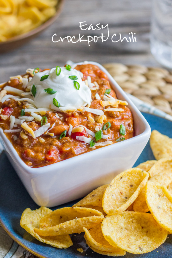 Easy Crockpot Chili in a bowl topped with shredded cheese and sour cream, sitting on a plate with Fritos. 