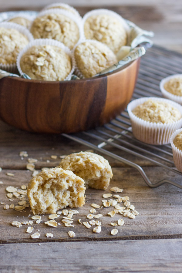 Healthy Applesauce Oat Muffin halved with oats sprinkled around it, and more muffins in a large bowl and on a cooling rack in the background.   