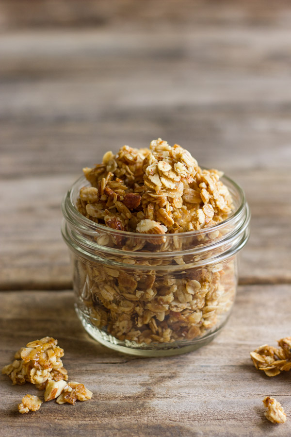 Homemade Coconut Oil Honey Almond Granola in a small glass jar with a few chunks spilled out next to it. 