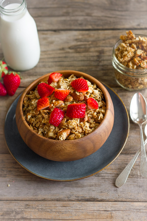 Homemade Coconut Oil Honey Almond Granola in a bowl with strawberry pieces on top, with a glass jar of milk and a small glass jar of the granola next to it. 