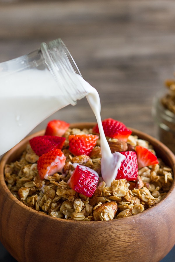 Homemade Coconut Oil Honey Almond Granola in a bowl with strawberry pieces on top, with a glass jar of milk being poured over it. 