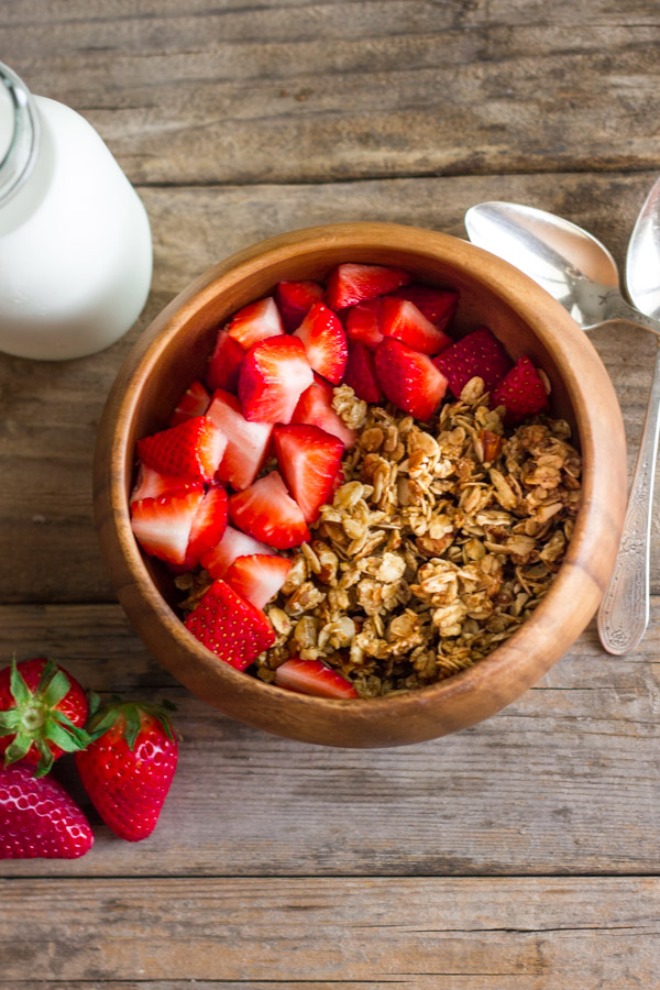 Homemade Coconut Oil Honey Almond Granola in a bowl with cut up strawberries, with a glass jar of milk, three whole strawberries and two spoons next to it. 
