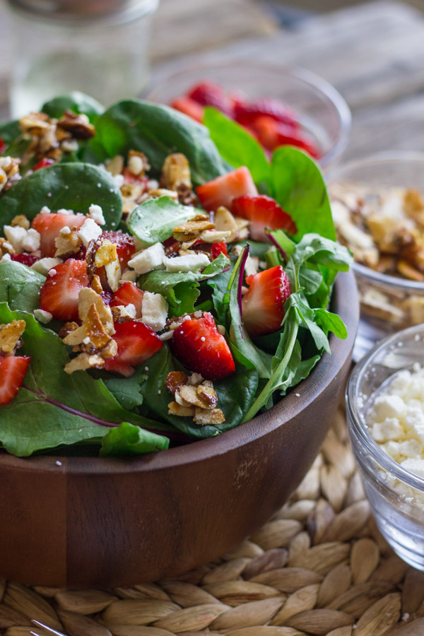 Strawberry and Spinach Salad with Almond Vinaigrette in a bowl, with a small glass dish of strawberries, a small glass dish of sugared almonds, and a small glass dish of Feta cheese next to it.  