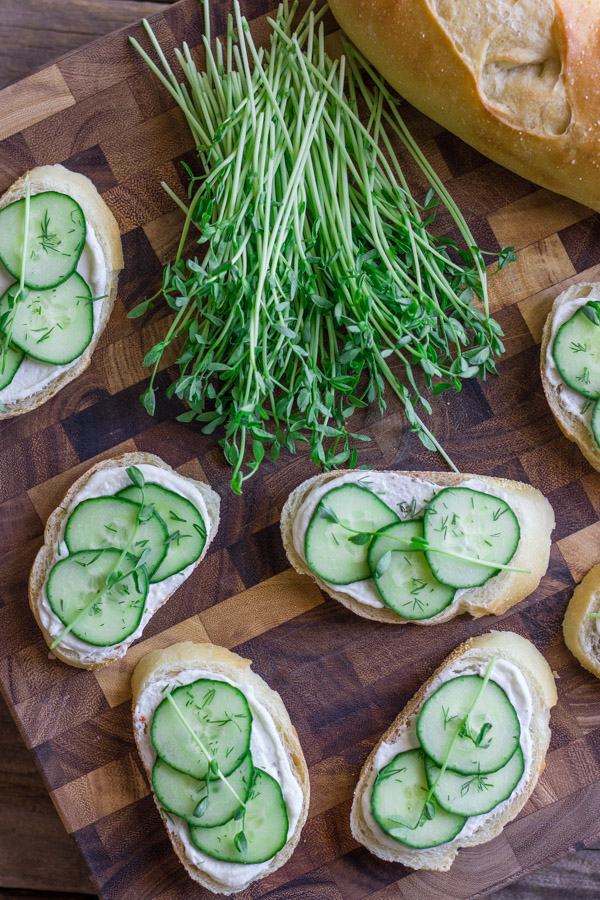 Cucumber Sandwiches With Whipped Goat Cheese on a cutting board with a bundle of pea shoots and a loaf of bread.  