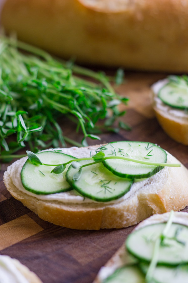 Cucumber Sandwiches With Whipped Goat Cheese on a cutting board with a bundle of pea shoots and a loaf of bread in the background.  