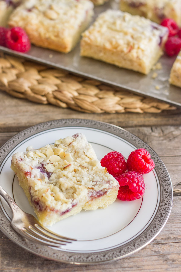 Raspberry Almond Crumb Bar square piece on a plate with fresh raspberries and a fork, with more Raspberry Almond Crumb Bar squares on a serving platter with fresh raspberries in the background.  