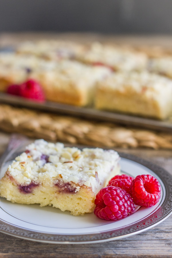 Raspberry Almond Crumb Bar square piece on a plate with fresh raspberries, with more Raspberry Almond Crumb Bar squares on a serving platter with fresh raspberries in the background.  
