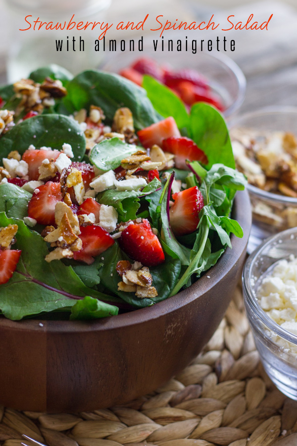 Strawberry and Spinach Salad with Almond Vinaigrette in a bowl, with a small glass dish of strawberries, a small glass dish of sugared almonds, and a small glass dish of Feta cheese next to it.  