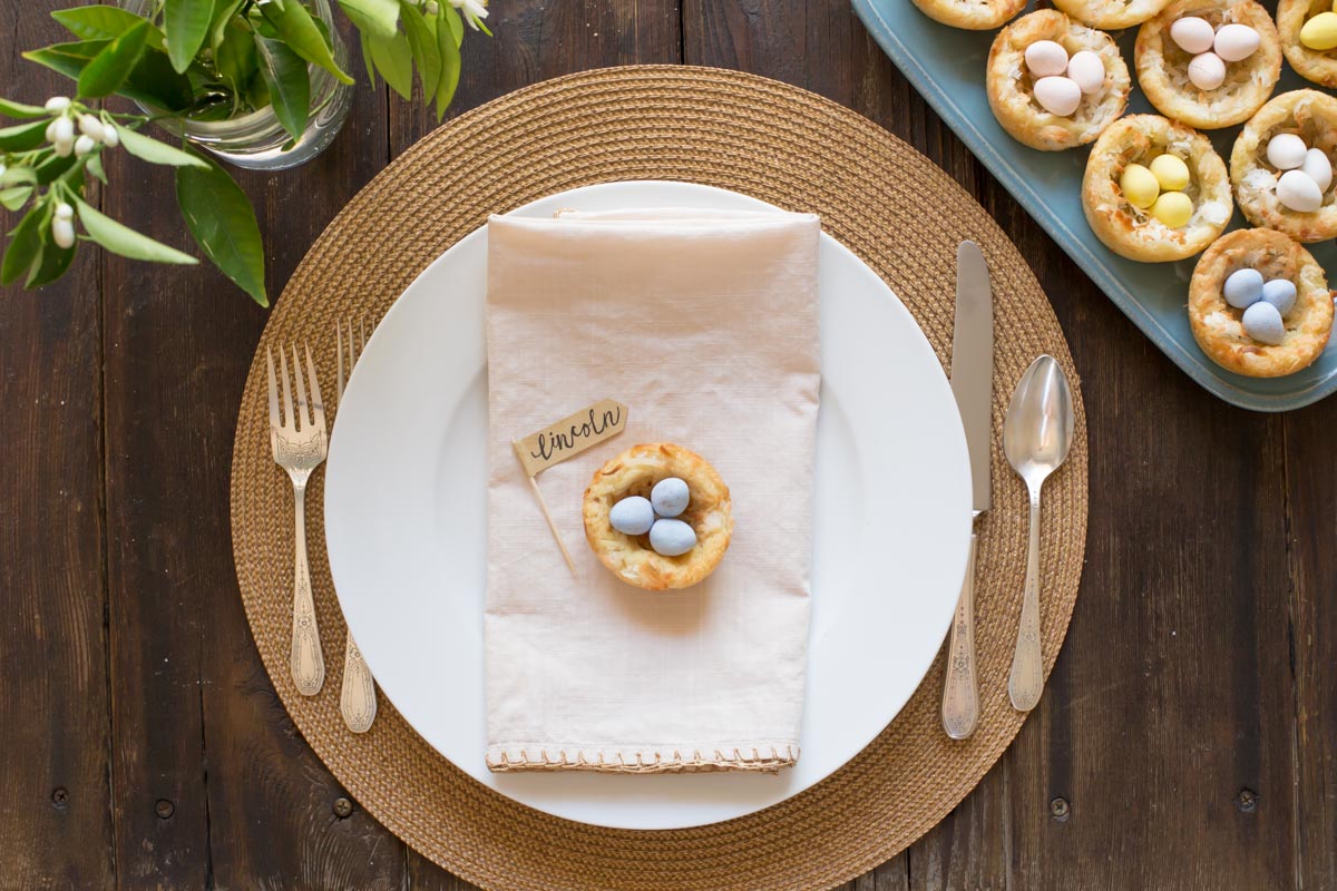 Sugar Cookie Easter Egg Nest with a name flag, sitting on top of a folded napkin on a place setting, with a vase of greenery and a platter of more Sugar Cookie Easter Egg Nests next to the place setting.  