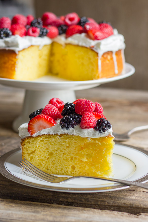 A slice of Easy Triple Berry Almond Poke Cake on a plate with a fork, and the rest of the cake on a cake stand in the background.  