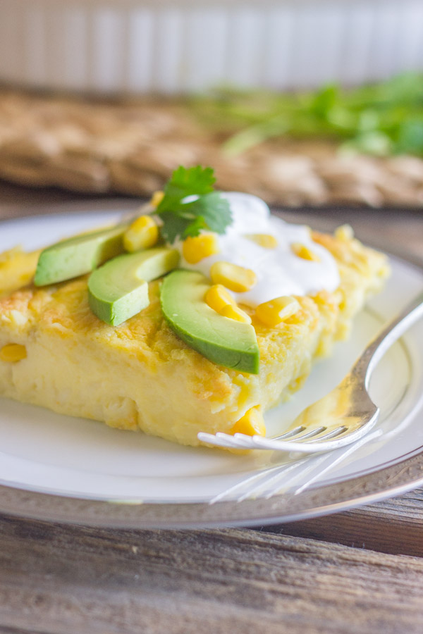 Green Chile Egg Bake Made With Greek Yogurt topped with avocado slices, sour cream, corn kernels and cilantro, on a plate with a fork.  