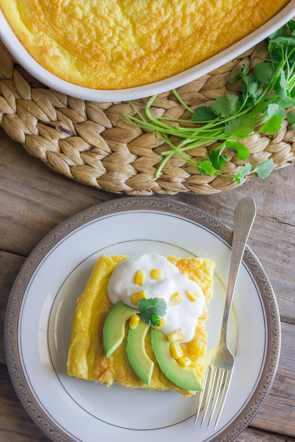 Green Chile Egg Bake Made With Greek Yogurt topped with avocado slices, sour cream, corn kernels and cilantro, on a plate with a fork, sitting next to the baking dish of the egg bake and a bundle of cilantro. 