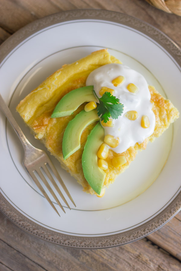 Green Chile Egg Bake Made With Greek Yogurt topped with avocado slices, sour cream, corn kernels and cilantro, on a plate with a fork.