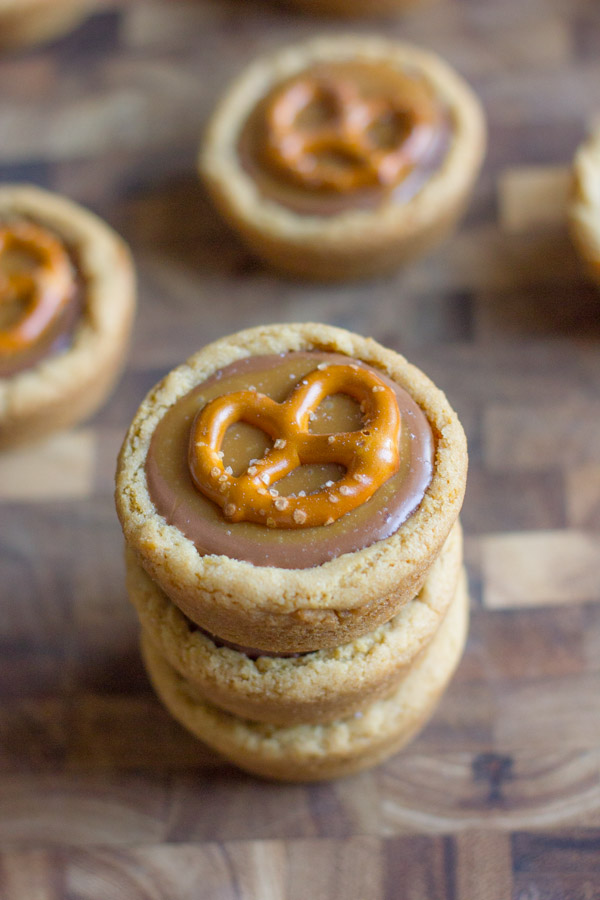 Peanut Butter Caramel Pretzel Cookie Cups stacked in a pile of three, surrounded by more Peanut Butter Caramel Pretzel Cookie Cups on a cutting board.  