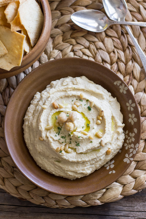 Simple Homemade Hummus in a bowl, topped with a drizzle of olive oil, dried parsley and pine nuts, with a bowl of pita chips and two spoons next to it.  