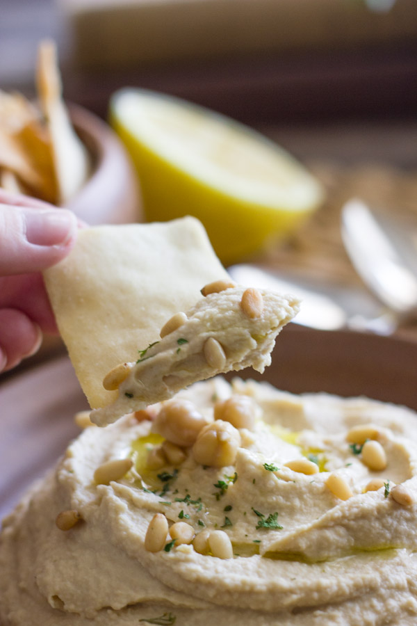 A pita chip dipped into a bowl of Simple Homemade Hummus that is topped with a drizzle of olive oil, dried parsley and pine nuts.