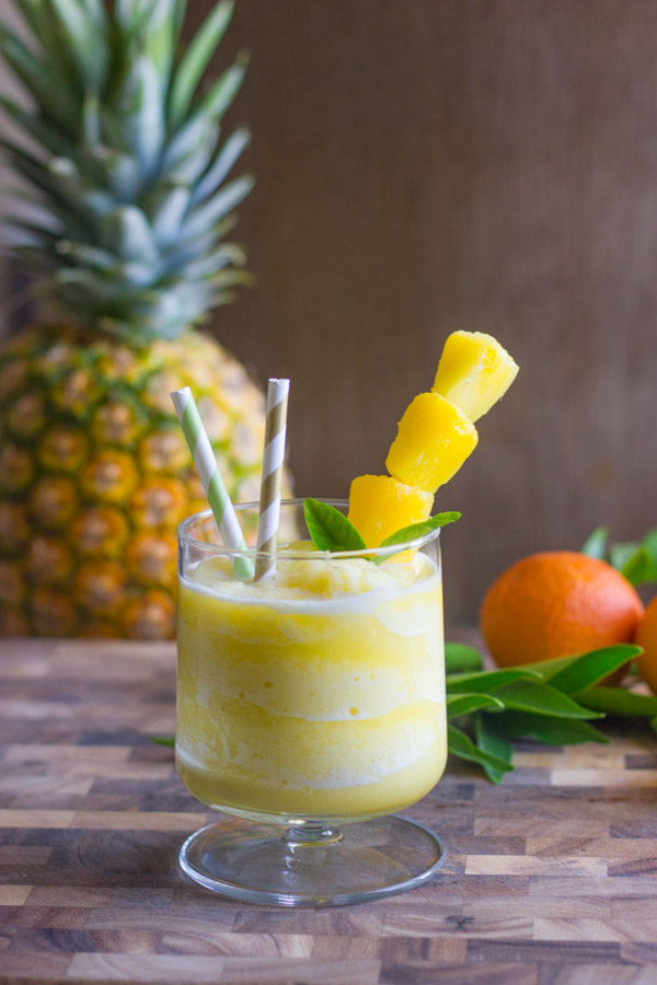 Skinny Pineapple Orange Slush in a glass with two straws and pineapple chunks on a stick, with a whole pineapple and an orange in the background.  