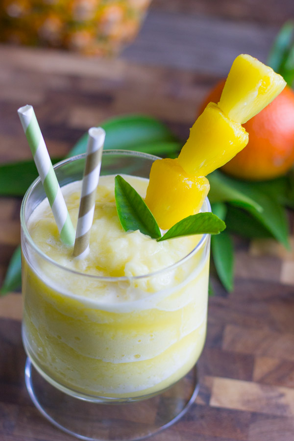 Skinny Pineapple Orange Slush in a glass with two straws and pineapple chunks on a stick, with an orange in the background.  