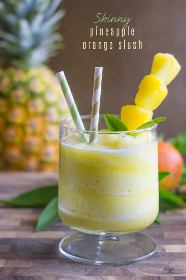 Skinny Pineapple Orange Slush in a glass with two straws and pineapple chunks on a stick, with a whole pineapple in the background.  