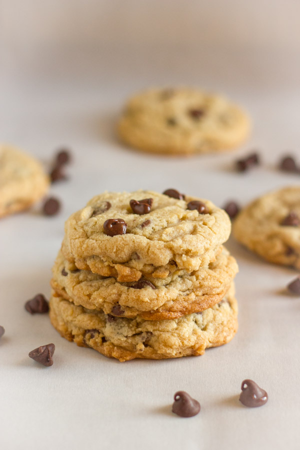 Super Soft Bakery Style Chocolate Chip Cookies stacked in a pile of three, with whole cookies and chocolate chips in the background.  
