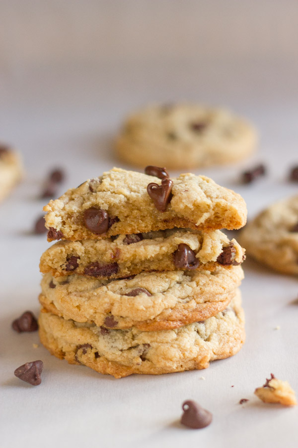 Two Super Soft Bakery Style Chocolate Chip Cookie halves stacked on top of two whole cookies, with chocolate chips and more cookies in the background.  