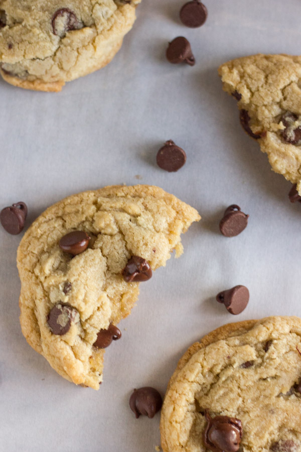 Super Soft Bakery Style Chocolate Chip Cookie broken in half, with chocolate chips and more cookies around it.  
